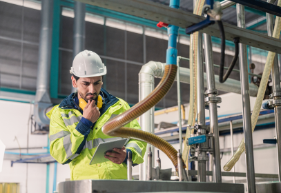 Man scratching his face and he appears to be thinking about preventive maintenance or repairs in a factory setting. He is looking a clip board and wearing a safety vest with a hard hat.