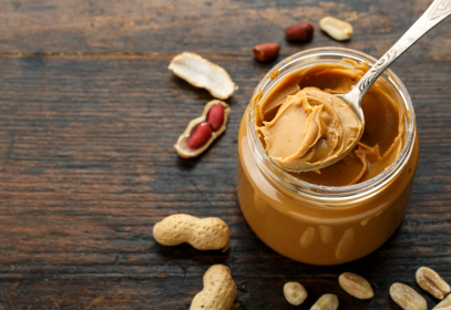 a jar of peanut butter with a spoon on a wooden table with peanuts scattered.