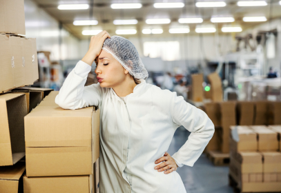 Woman holding her hand to her head wearing a hair net in a food industry factory with corrugated boxes all around her.