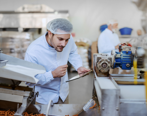 Man wearing a hairnet and food factory uniform inspecting a machine while holding an electronic device.