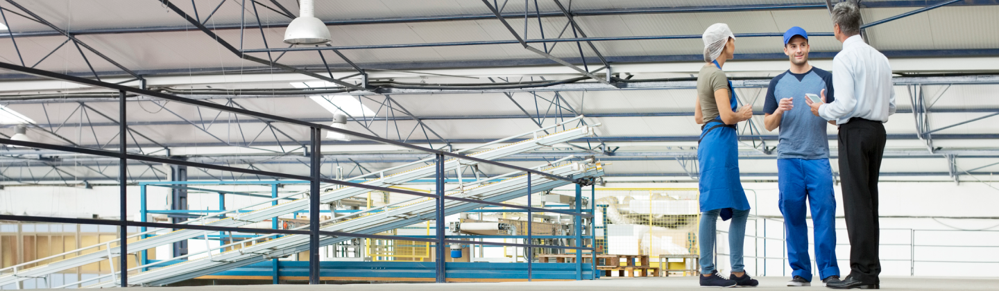two men and a woman standing and talking inside and industrial building wearing work uniforms.