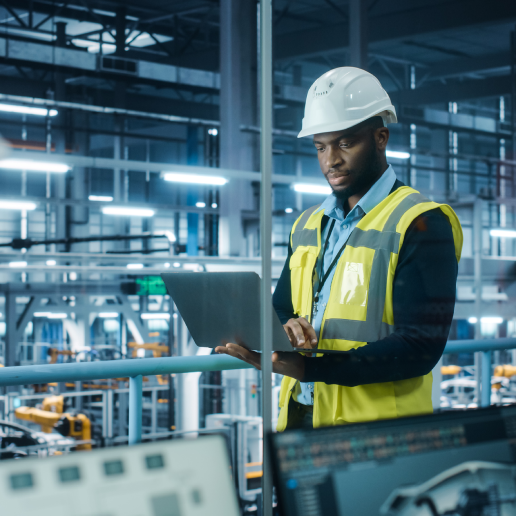 Man wearing a safety vest and helmet in a factory and looking down at a computer.